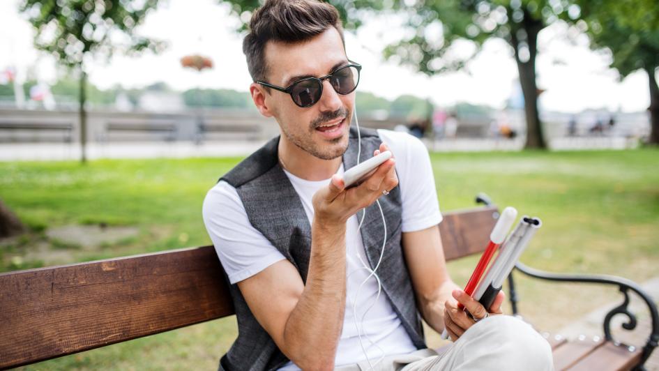 Young blind man with smartphone sitting on bench in park in city, calling