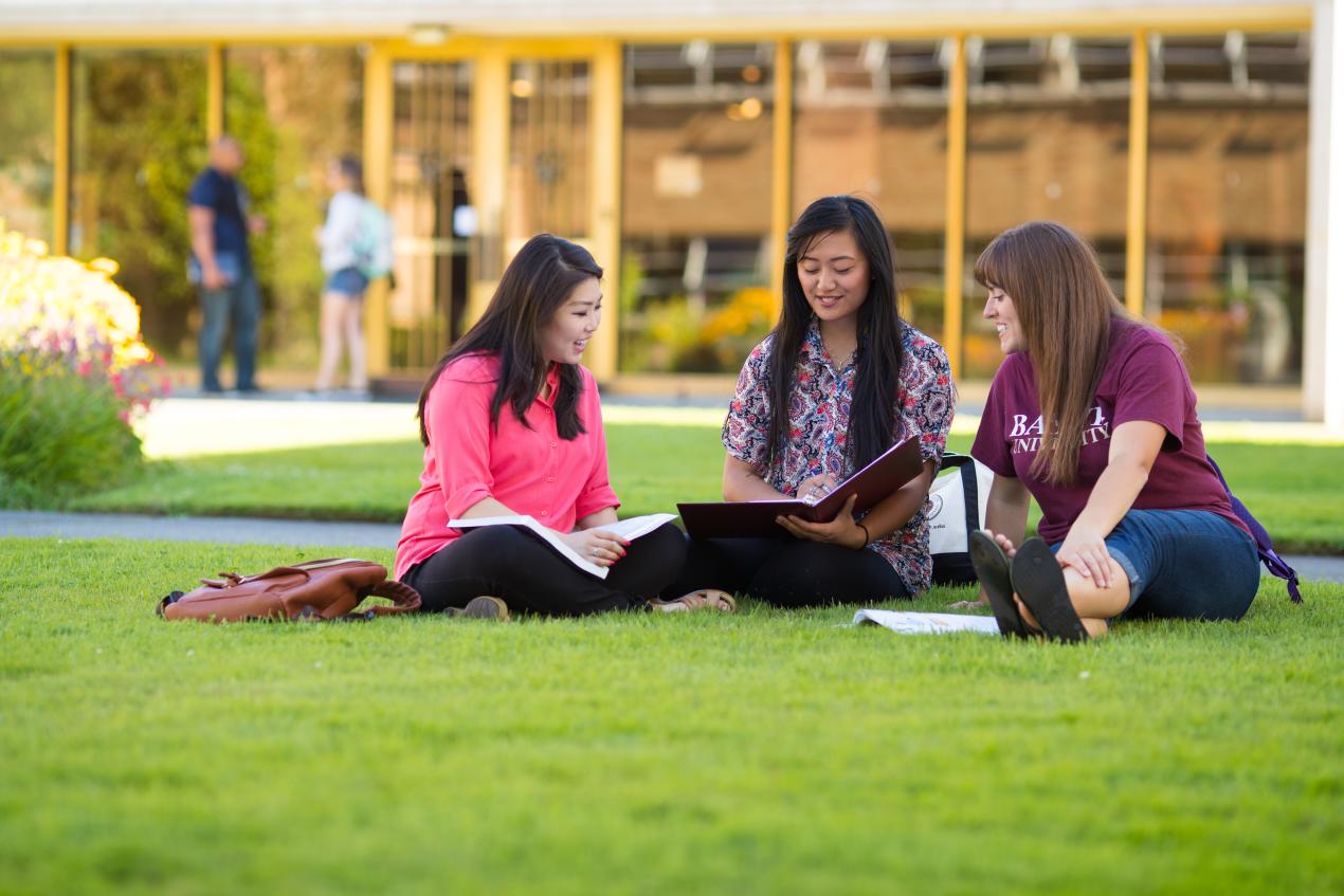 three students sitting on lawn looking at notebooks